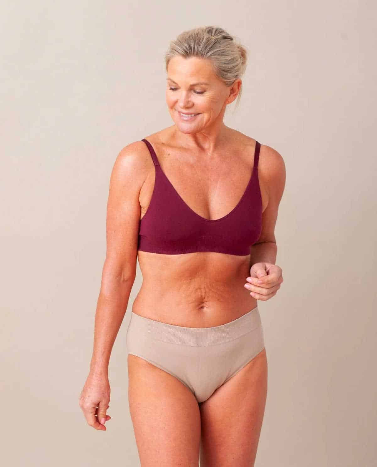 Smiling grey haired person wearing a burgundy bra and oatmeal coloured knickers.