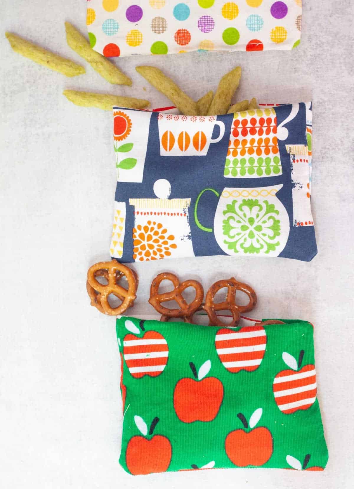 Three reusable snack bags in colourful patterned fabric, with pretzels and crisps spilling out of them.