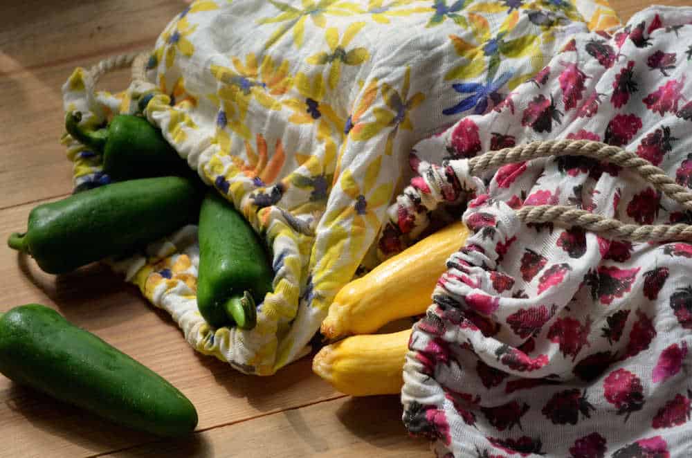 Two floral zero-waste reusable produce bags filled with green peppers and yellow squashes.