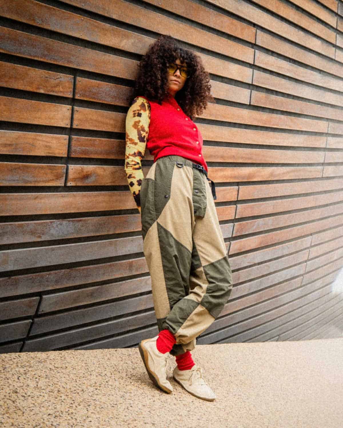 Stylish person standing against a slatted wooden wall, wearing fashionable clothes and white trainers from sustainable brand Vivobarefoot.