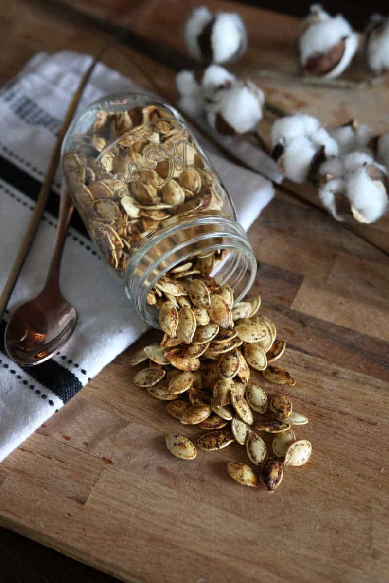 Roasted pumpkin seeds spilling out of a jar onto a wooden chopping board.