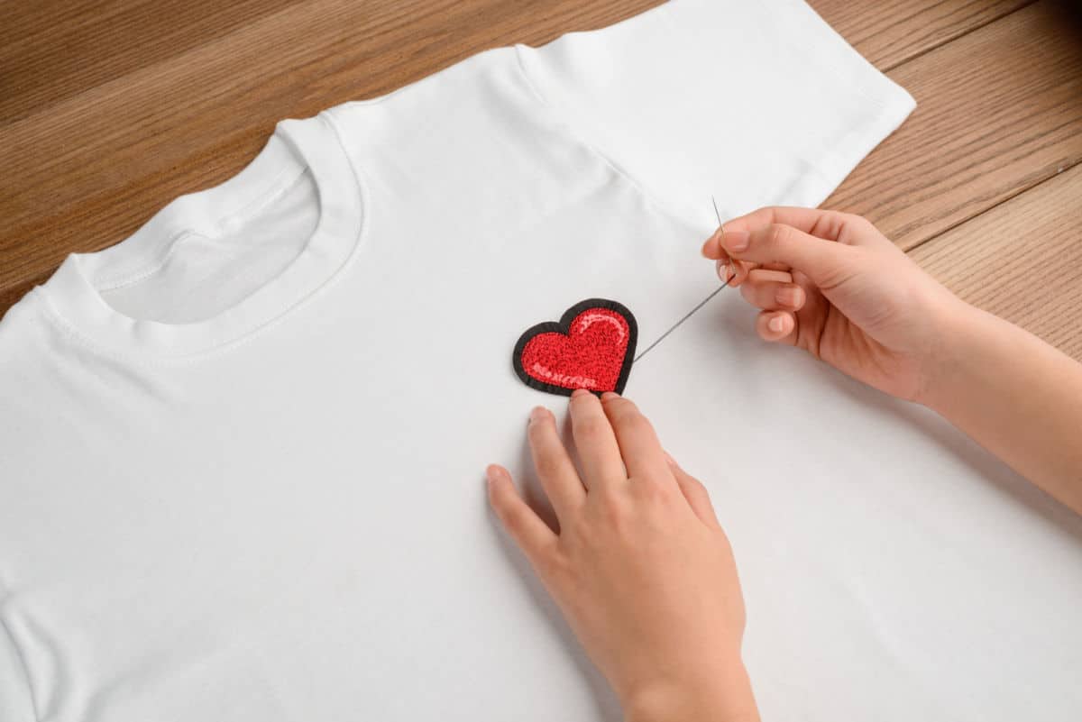 Person sewing a red heart patch onto a white t-shirt.